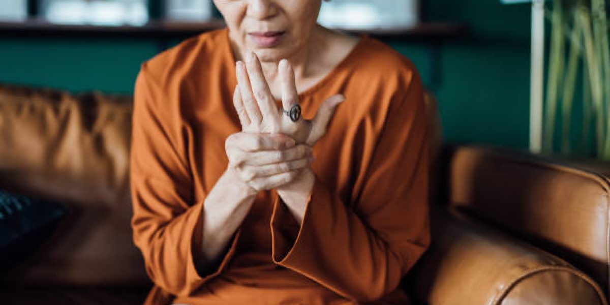 Senior Asian woman rubbing her hands in discomfort, suffering from arthritis in her hand while sitting on sofa at home. Elderly and health issues concept